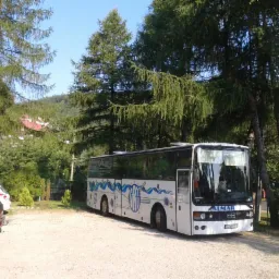 Biały autokar z niebieskimi zdobieniami marki Almar zaparkowany na żwirowym parkingu, otoczony zielenią drzew i krzewów w słoneczny dzień. Widoczne zaparkowane samochody osobowe.