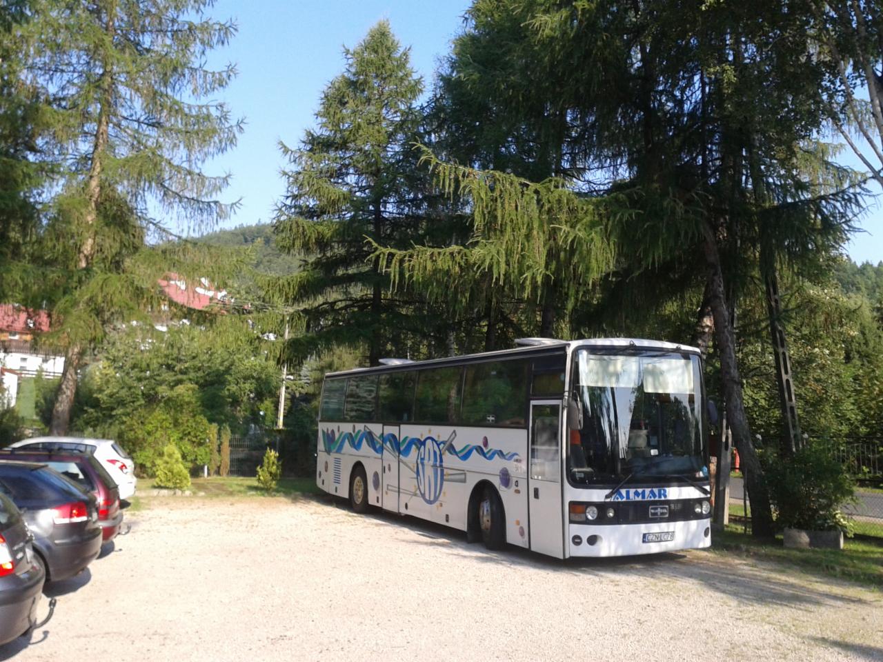 Biały autokar z niebieskimi zdobieniami marki Almar zaparkowany na żwirowym parkingu, otoczony zielenią drzew i krzewów w słoneczny dzień. Widoczne zaparkowane samochody osobowe.