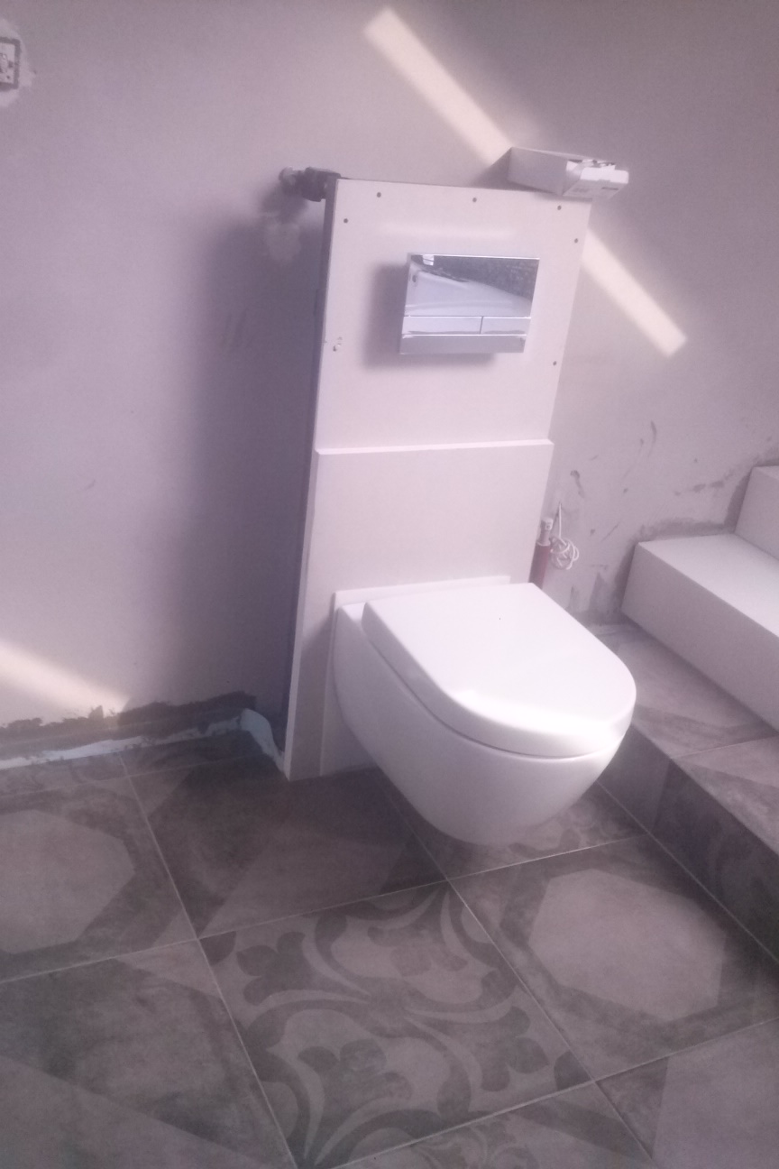 Biała toaleta podwieszana na tle ściany, z widocznymi szarymi płytkami podłogowymi z wzorem. Elementy instalacji sanitarnej widoczne na ścianie.