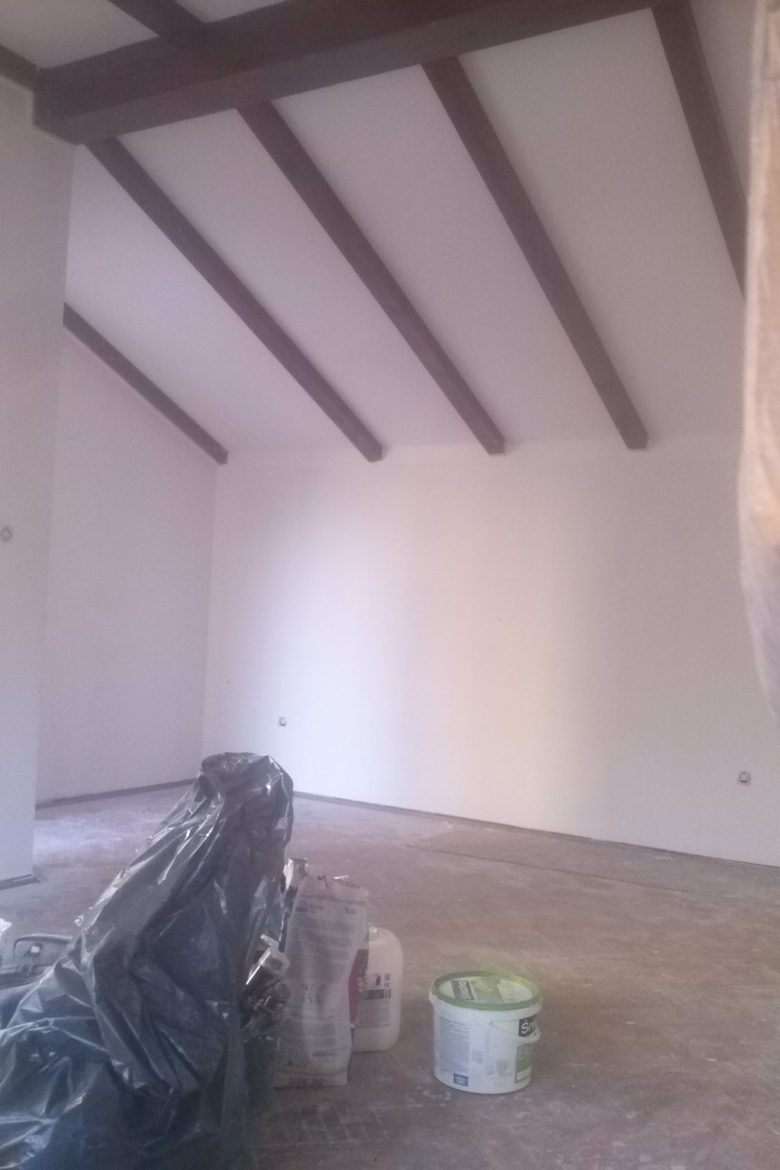 Wnętrze po malowaniu z drewnianymi belkami stropowymi. Na podłodze wiadro farby i zakryte folią materiały budowlane. Ściany świeżo pomalowane na biało.