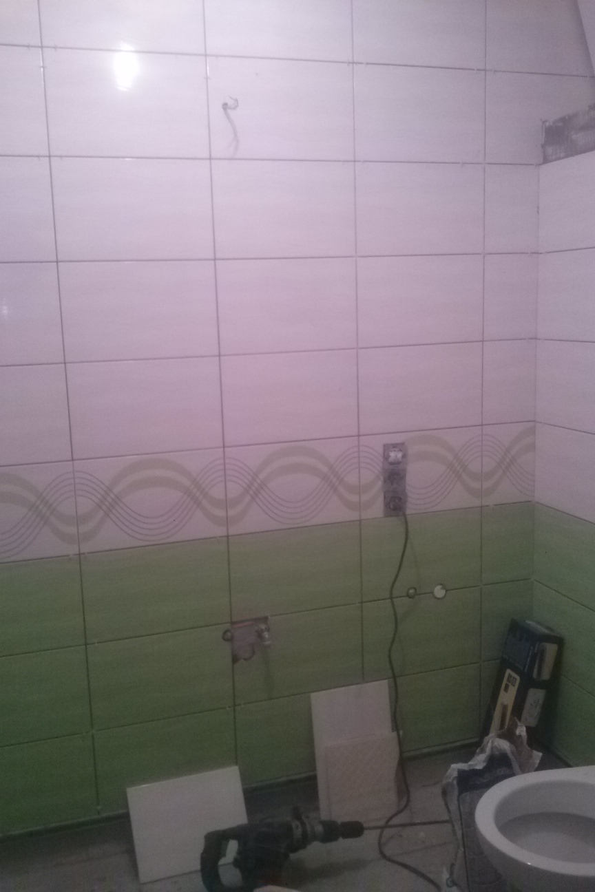 Łazienka w trakcie remontu: biało-zielone płytki ścienne z dekoracyjnym pasem, widoczne narzędzia (wiertarka) i niedokończone elementy instalacji elektrycznej.