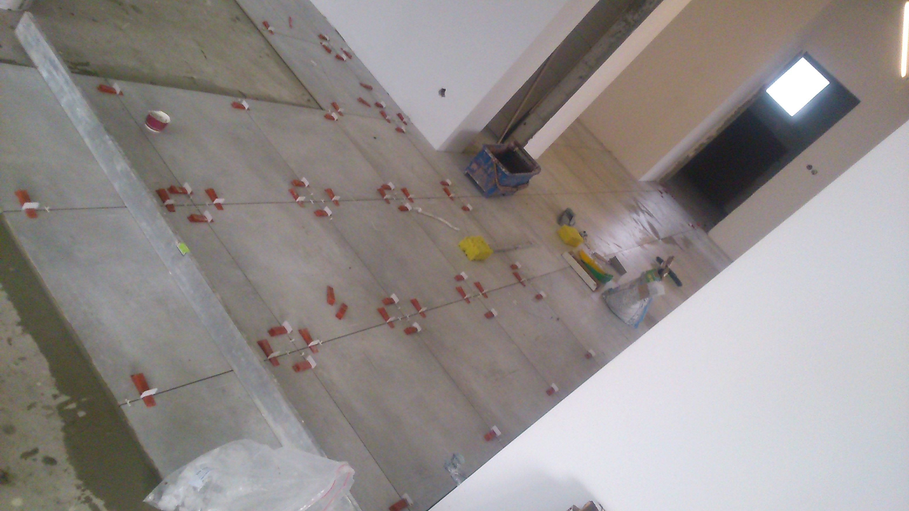 Wnętrze w trakcie prac: szare płytki podłogowe z czerwonymi krzyżykami dystansowymi, poziomica, wiadro i narzędzia glazurnicze. Widok na dalszą część pomieszczenia.