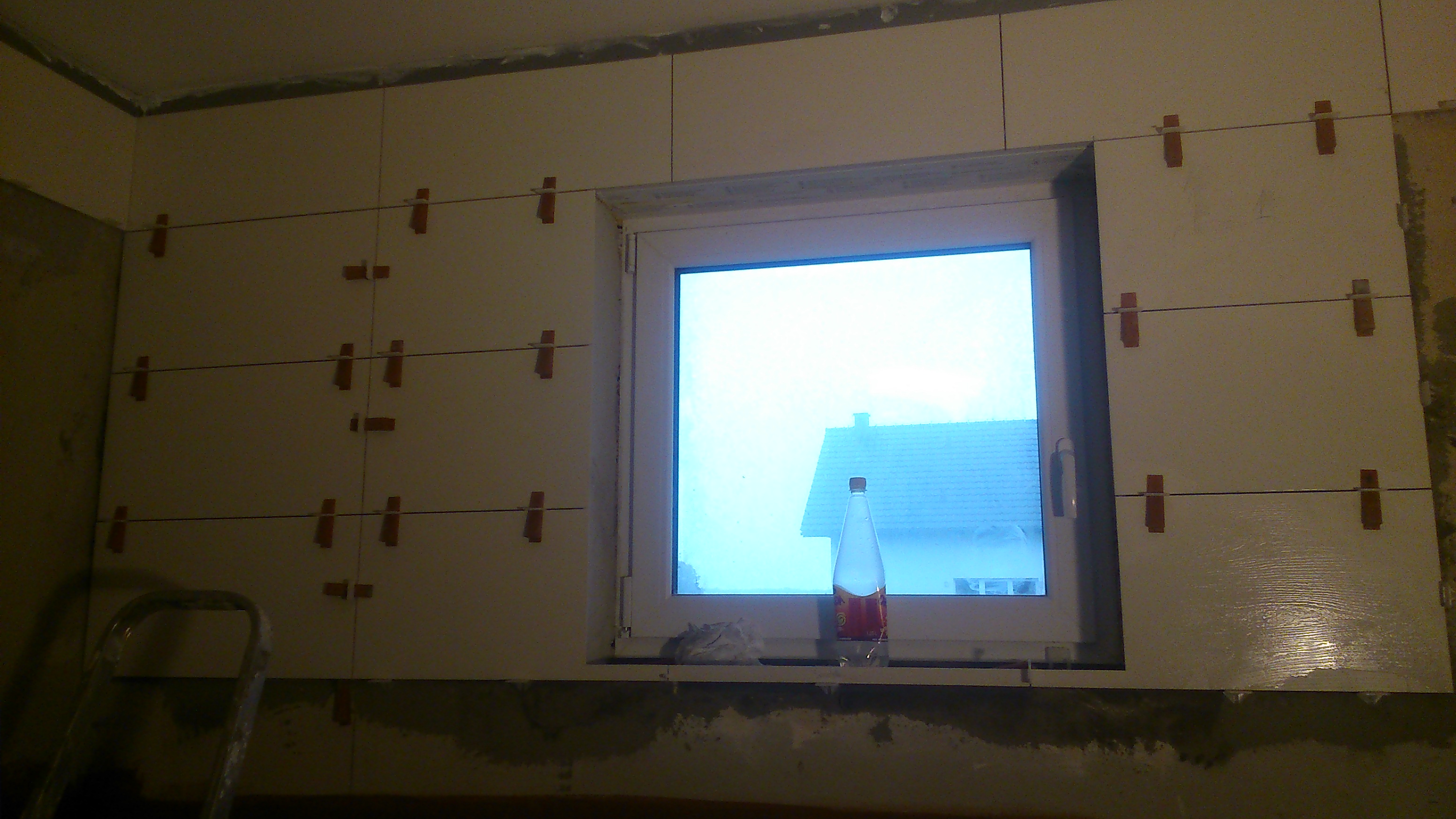 Ściana z oknem w trakcie układania płytek, widoczne krzyżyki dystansowe i pozioma linia. Na parapecie butelka. Dolna część ściany przygotowana pod płytki.
