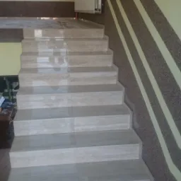 Wykończone schody wewnętrzne z jasnymi płytkami, ściany w odcieniach beżu i brązu z dekoracyjnymi pasami. Widoczne narzędzia i butelka na dole schodów.