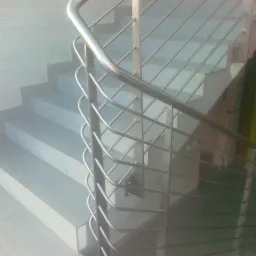 Metalowa balustrada schodowa z wypełnieniem z cienkich prętów, widoczna z boku, oświetlona naturalnym światłem, z delikatnym odbiciem na stopniach.