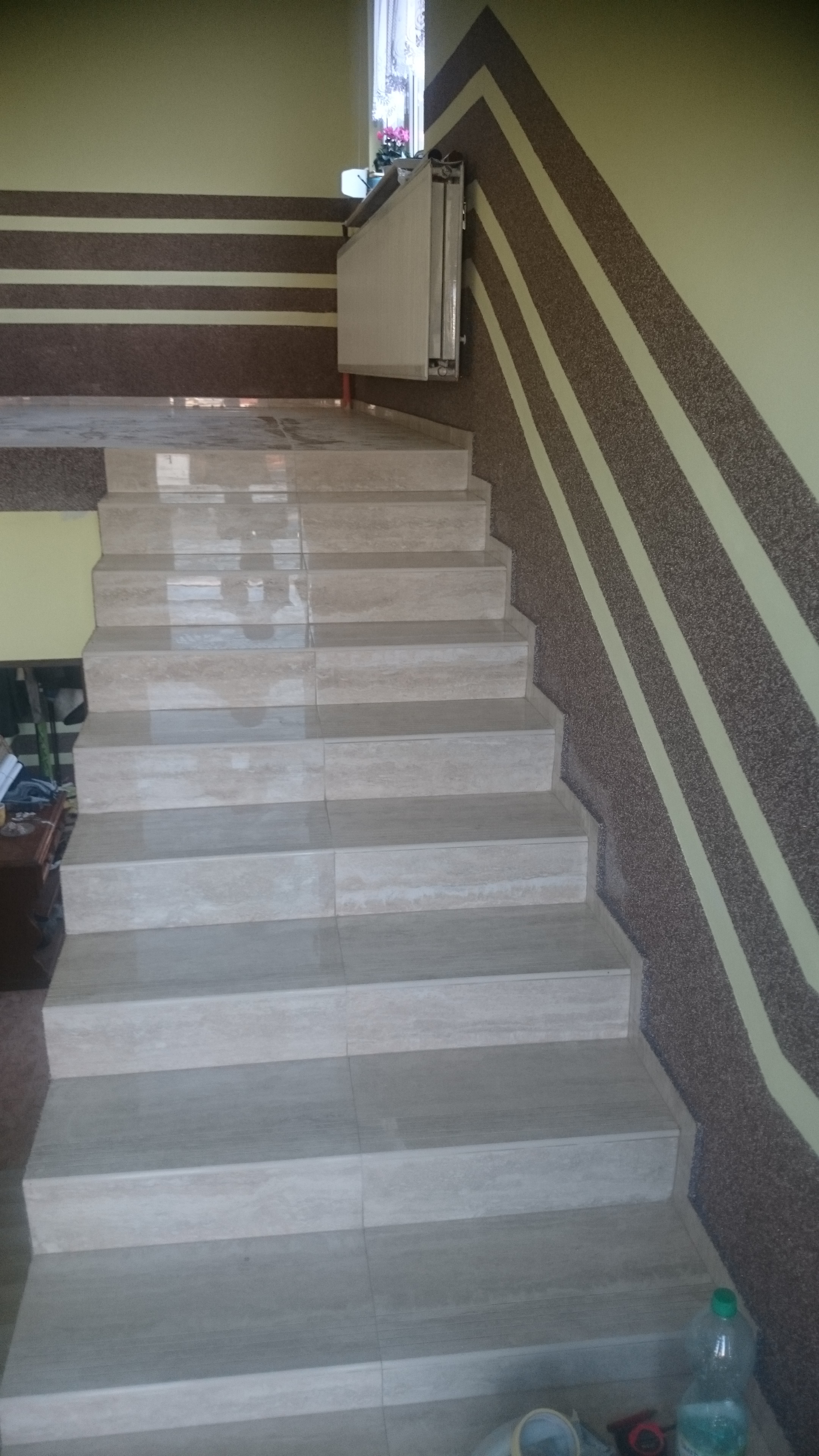 Wykończone schody wewnętrzne z jasnymi płytkami, ściany w odcieniach beżu i brązu z dekoracyjnymi pasami. Widoczne narzędzia i butelka na dole schodów.