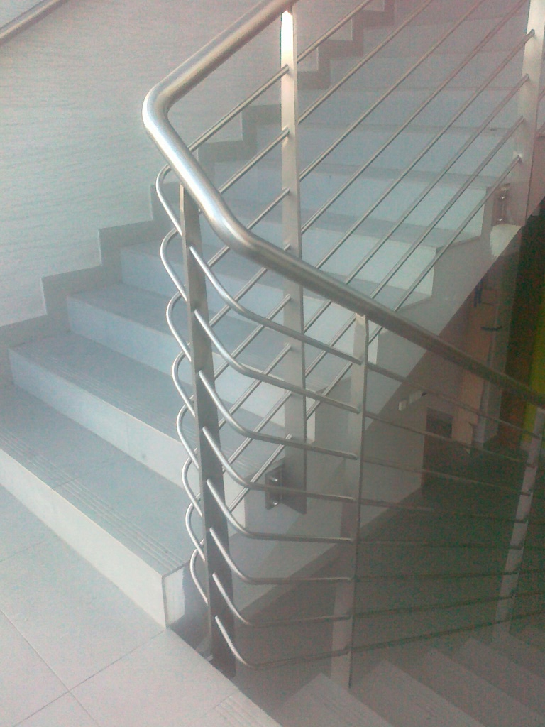 Metalowa balustrada schodowa z wypełnieniem z cienkich prętów, widoczna z boku, oświetlona naturalnym światłem, z delikatnym odbiciem na stopniach.