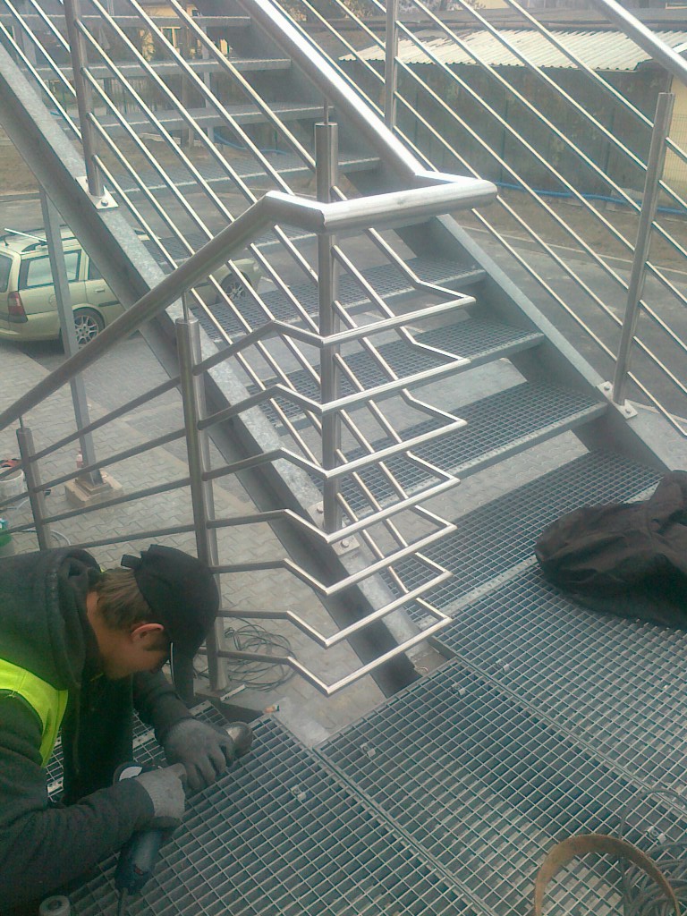 Montaż metalowej balustrady schodowej zewnętrznej przez pracownika w kamizelce, szlifowanie spawów na kratownicy. Widoczne narzędzia i elementy konstrukcji.