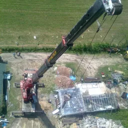 Czerwony dźwig teleskopowy w trakcie pracy na placu budowy z gruzem i elementami konstrukcyjnymi, widok z góry na zielone tereny.