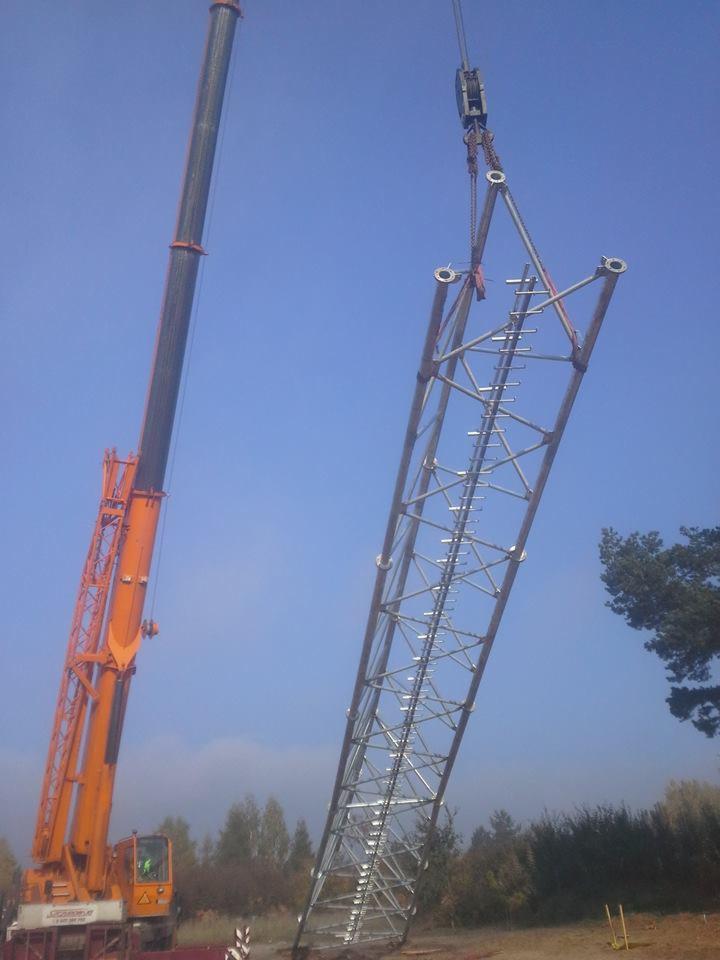 Pomarańczowy dźwig podnosi metalową konstrukcję wieży telekomunikacyjnej na tle błękitnego nieba. Widoczne drzewa w oddali.