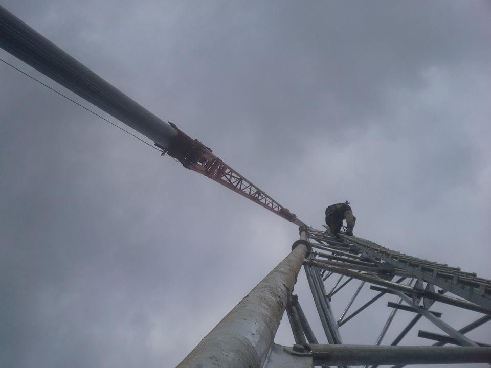 Pracownik wspina się po konstrukcji dźwigu teleskopowego na tle pochmurnego nieba, widok z dołu, w kadrze fragment liny dźwigu.