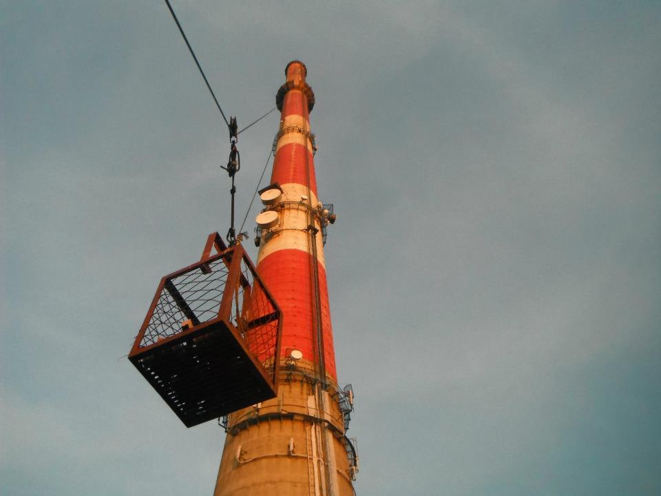 Metalowy kosz podwieszony na linie, na tle wysokiego komina z czerwono-białymi pasami i pochmurnego nieba. Ujęcie z dołu, podkreślające wysokość konstrukcji.