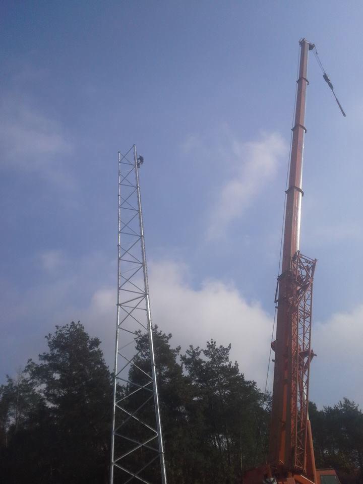 Montaż wieży telekomunikacyjnej z użyciem dźwigu. Pracownik na wysokości w trakcie instalacji, widok z dołu na tle błękitnego nieba z chmurami i drzew.