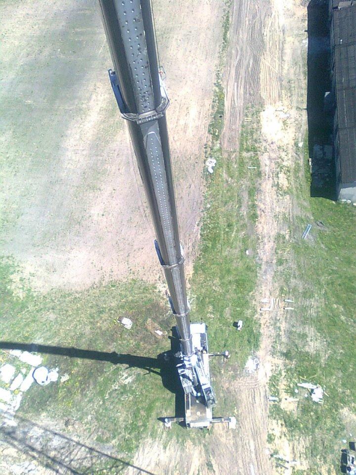 Widok z góry na wysięgnik dźwigu teleskopowego na trawiastym terenie z budynkiem w tle. Długi cień rzucany przez ramię dźwigu.