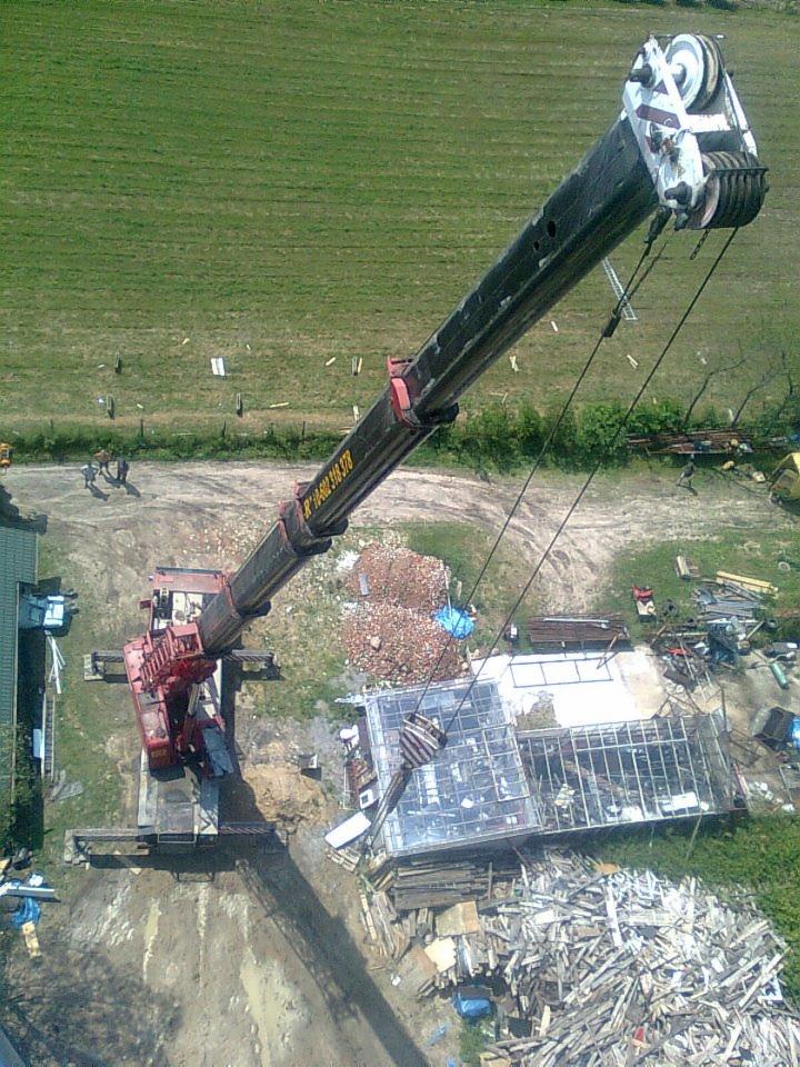 Czerwony dźwig teleskopowy w trakcie pracy na placu budowy z gruzem i elementami konstrukcyjnymi, widok z góry na zielone tereny.