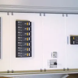 Rozdzielnica elektryczna z zamontowanymi bezpiecznikami i wyłącznikiem głównym, widok z bliska na białą płytę montażową i zainstalowane komponenty.