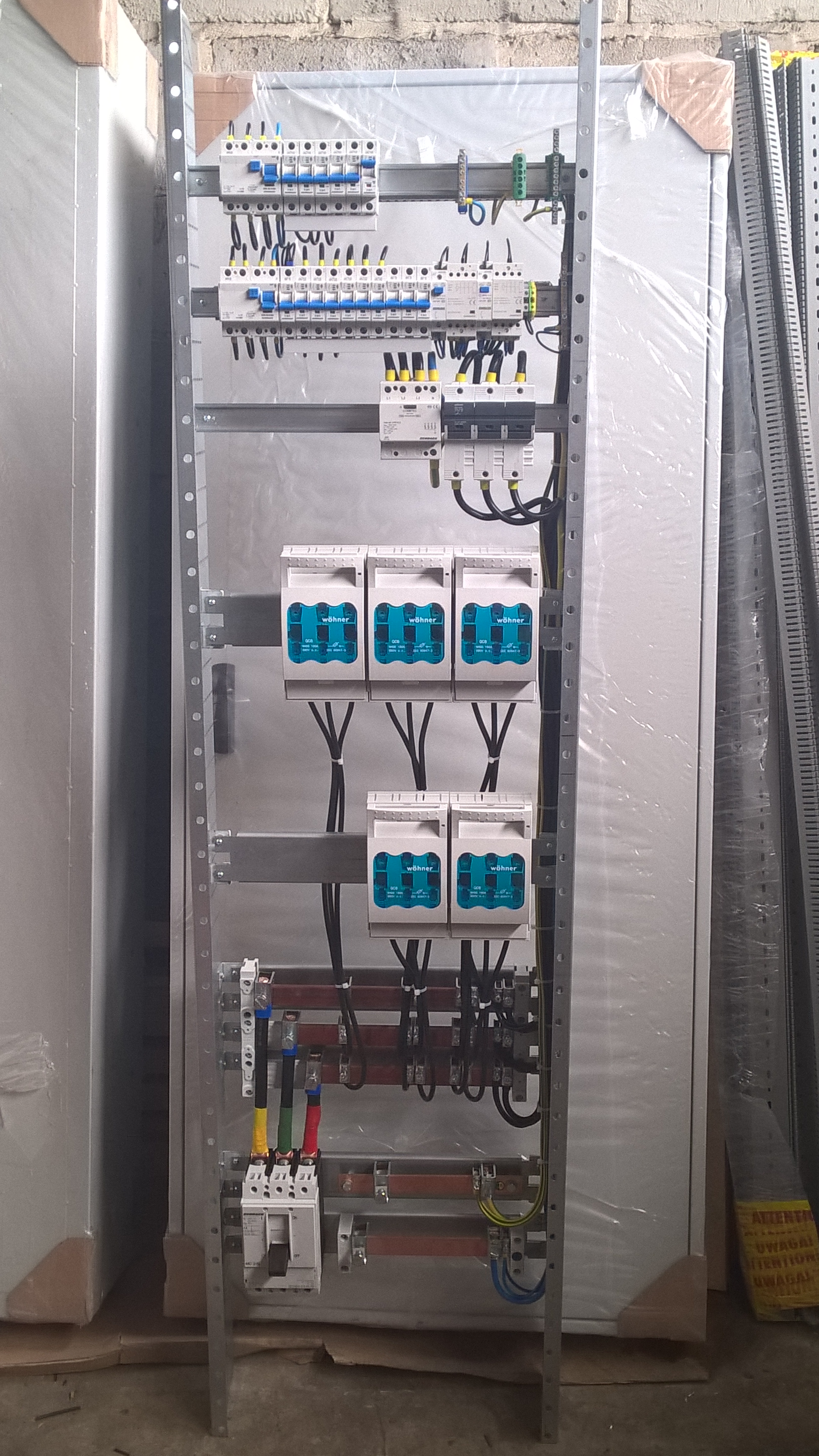 Rozdzielnica elektryczna w trakcie montażu, widoczne zabezpieczenia, przewody i szyny. Całość w metalowej obudowie, jeszcze częściowo w folii ochronnej.