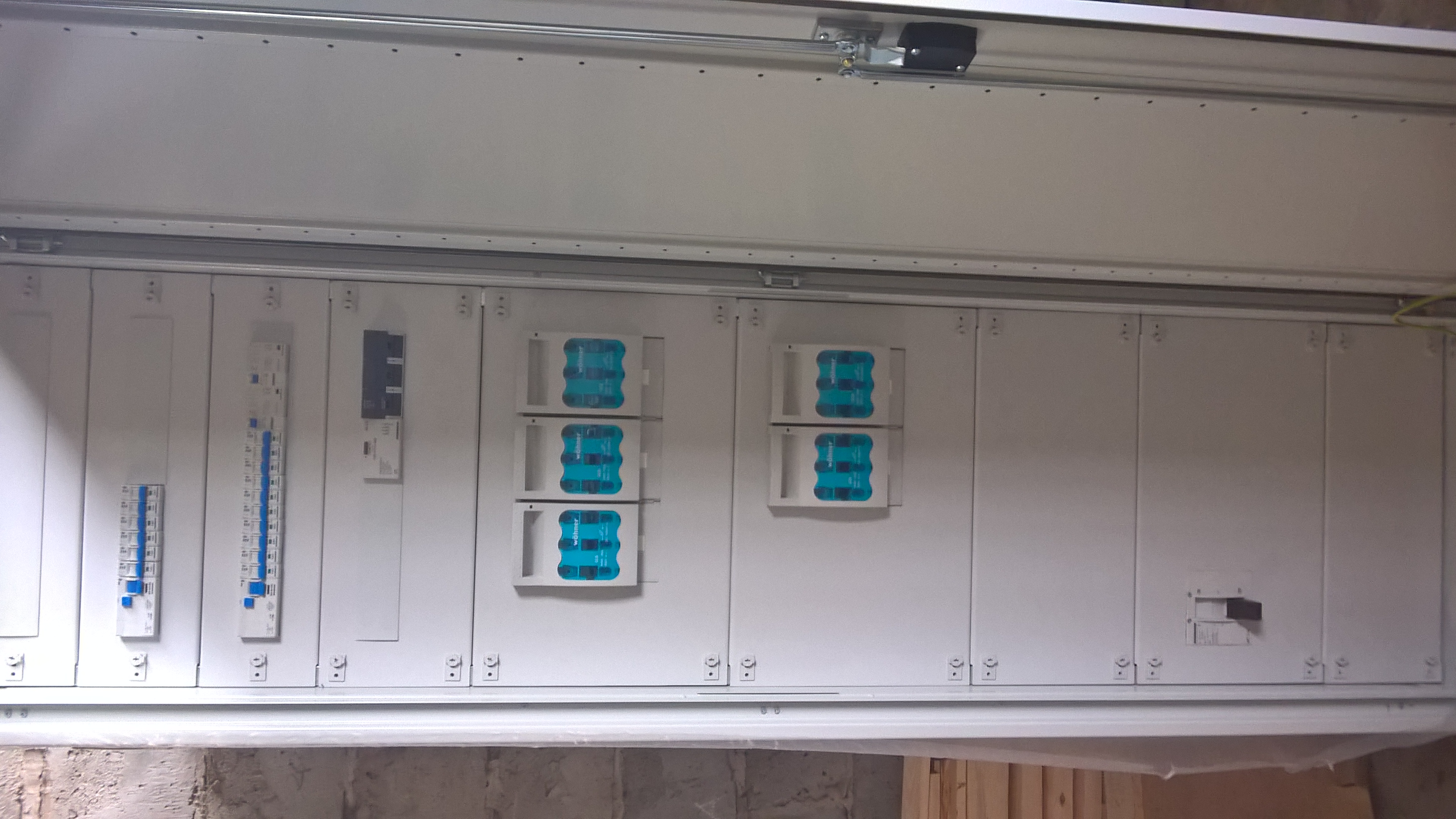 Rozdzielnia elektryczna z zabezpieczeniami, widok z dołu, w tle fragment ściany. Widoczne bezpieczniki i inne elementy instalacji wewnątrz obudowy.