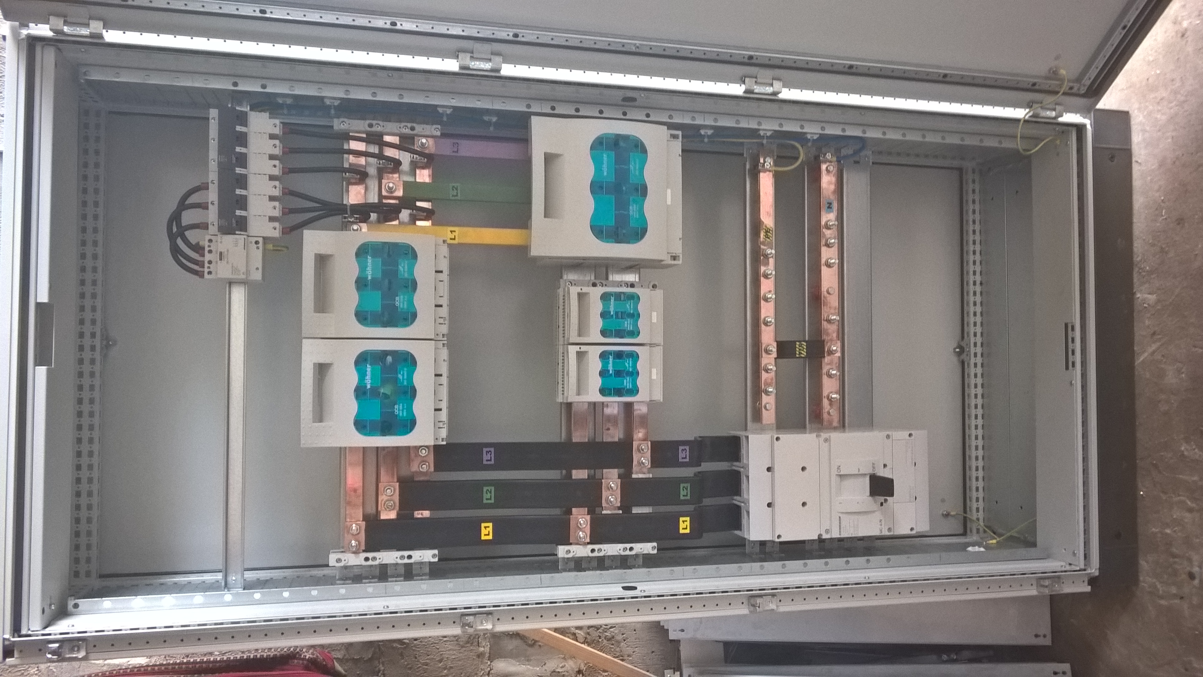 Otwarta szafa elektryczna z zainstalowanymi bezpiecznikami, przewodami i szynami zbiorczymi. Widoczne oznaczenia faz L1, L2, L3. Przewody uporządkowane.