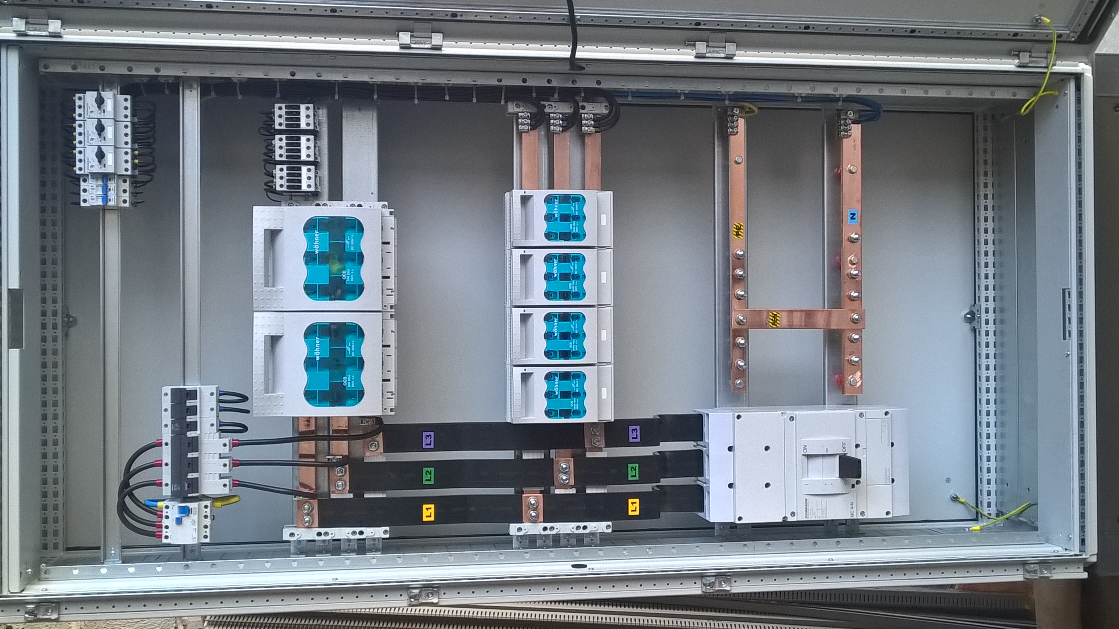 Rozdzielnia elektryczna w szafie sterowniczej z widocznymi bezpiecznikami, szynami zbiorczymi i przewodami oznaczonymi kolorami, zamontowana wewnątrz szarej obudowy.