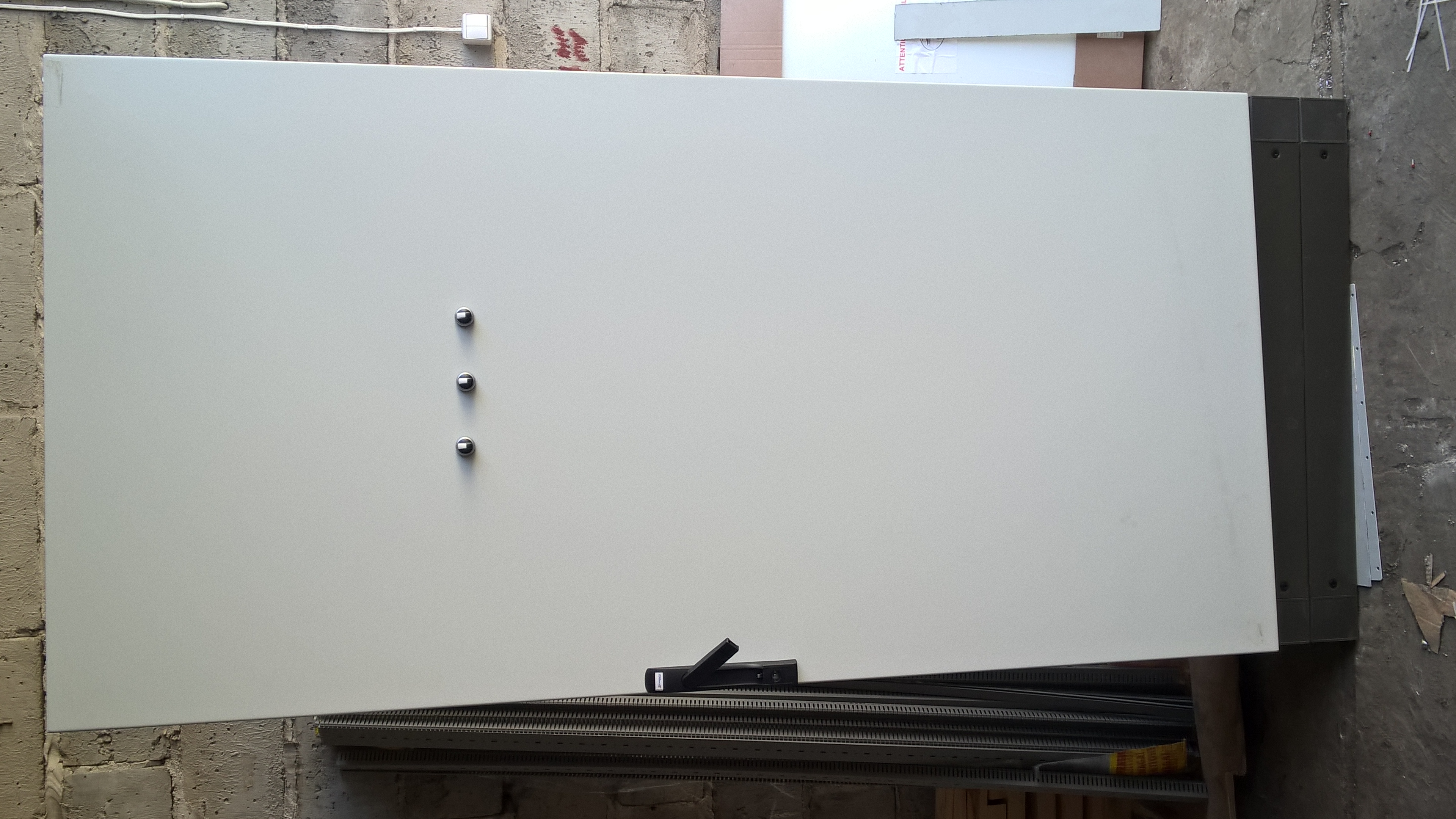 Rozdzielnica elektryczna z trzema metalowymi zatrzaskami, uchylona, widoczne szyny DIN wewnątrz, na tle betonowej ściany i podłogi.