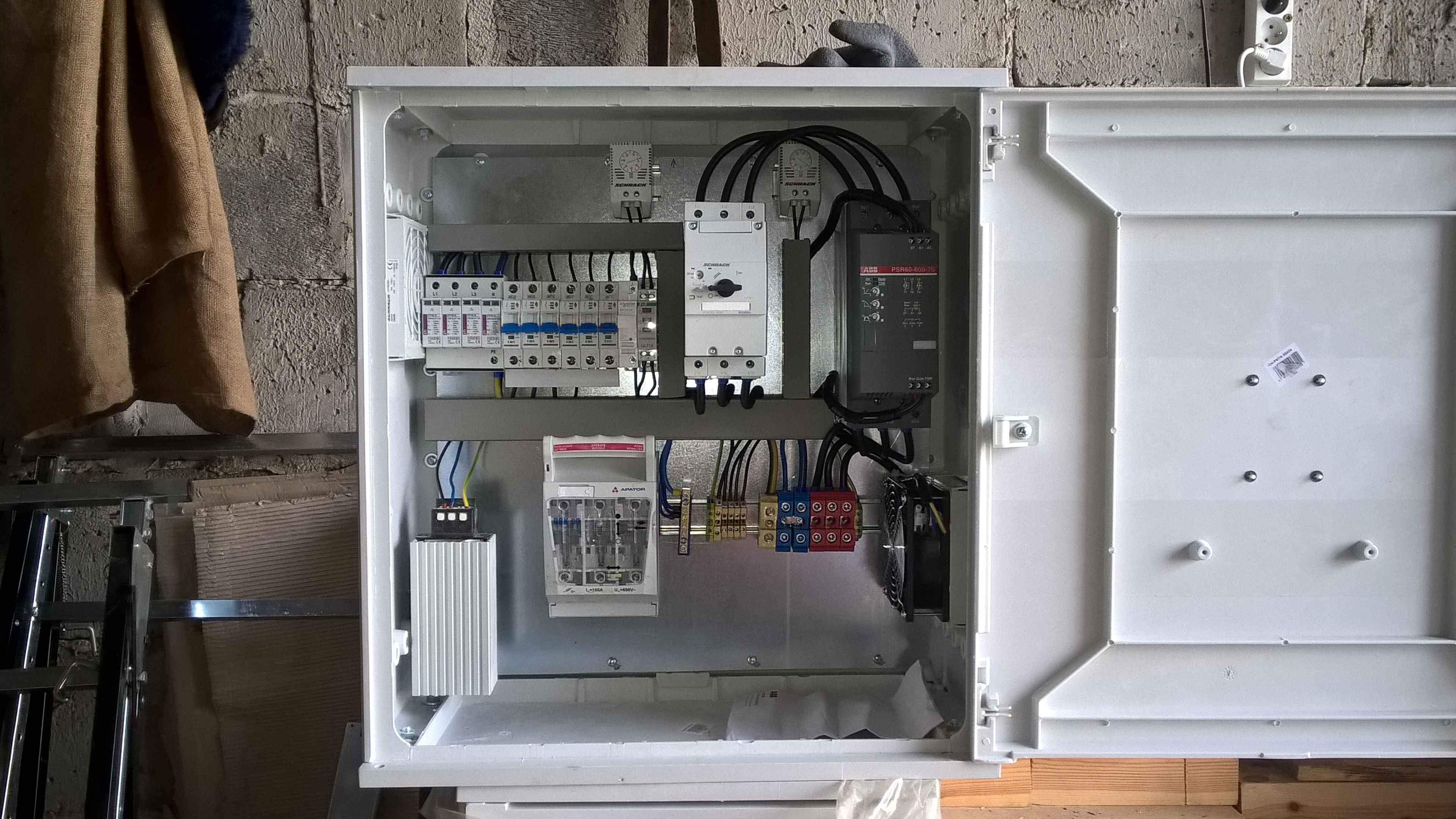 Otwarta skrzynka elektryczna z zainstalowanymi bezpiecznikami, stycznikami i przewodami, widok z bliska na szarą ścianę z cegieł w tle.