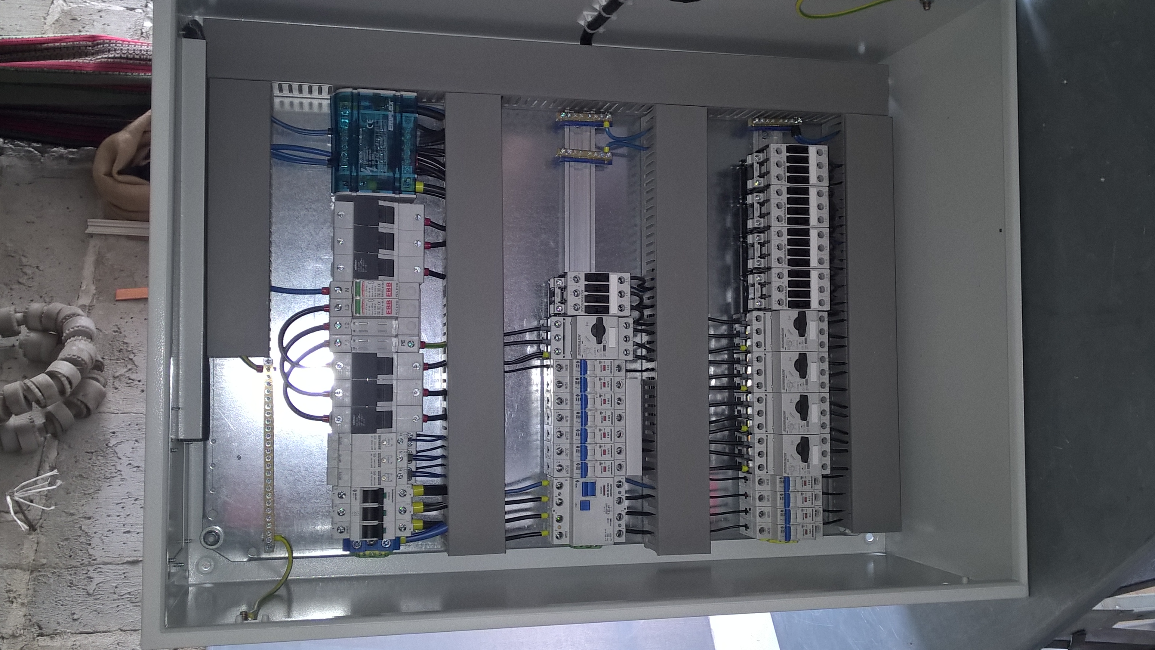 Wnętrze rozdzielnicy elektrycznej z zamontowanymi bezpiecznikami i okablowaniem, widok z góry. Szara obudowa, elementy zabezpieczeń w kolorach białym, niebieskim i czarnym.