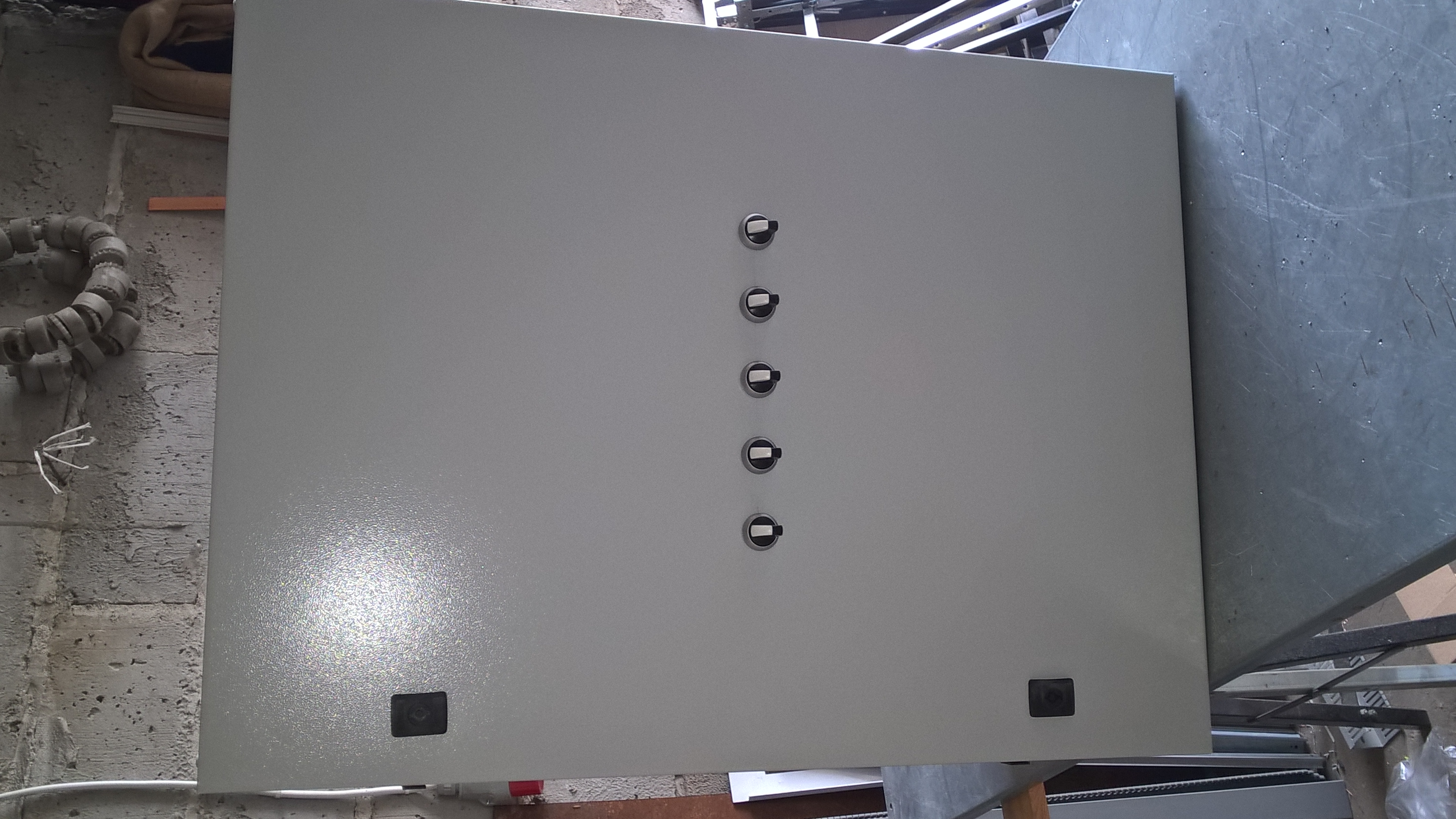 Szara, metalowa obudowa rozdzielnicy elektrycznej z rzędem przełączników i dwoma czarnymi elementami montażowymi w dolnej części, na tle surowej ściany i metalowej konstrukcji.