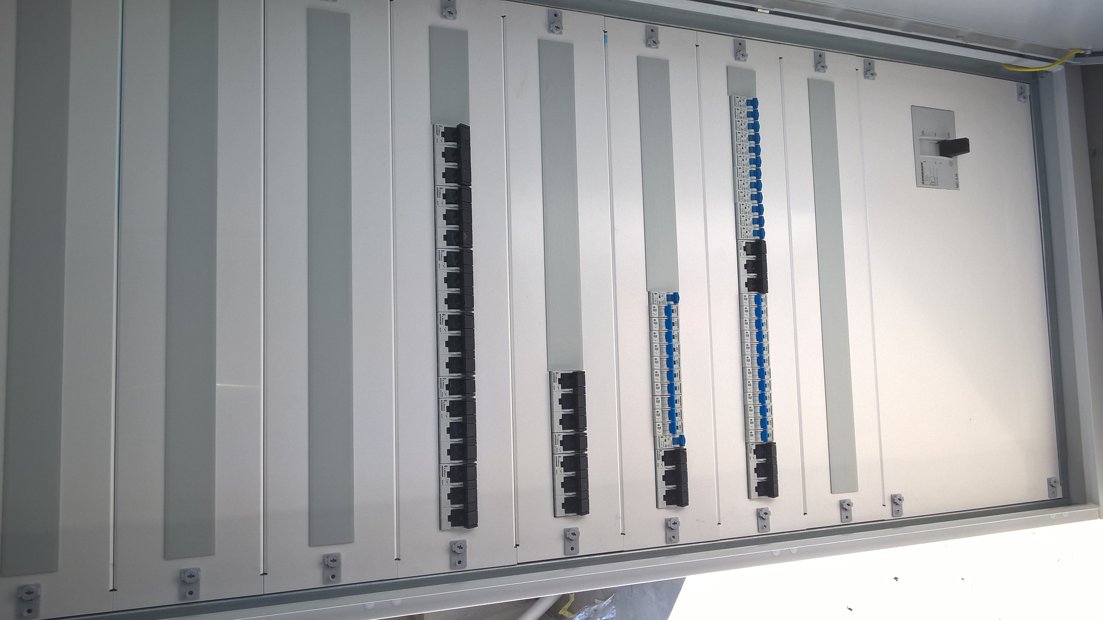 Szara, metalowa skrzynka elektryczna z zamontowanymi bezpiecznikami i wyłącznikiem. Widoczne listwy montażowe i elementy mocujące.