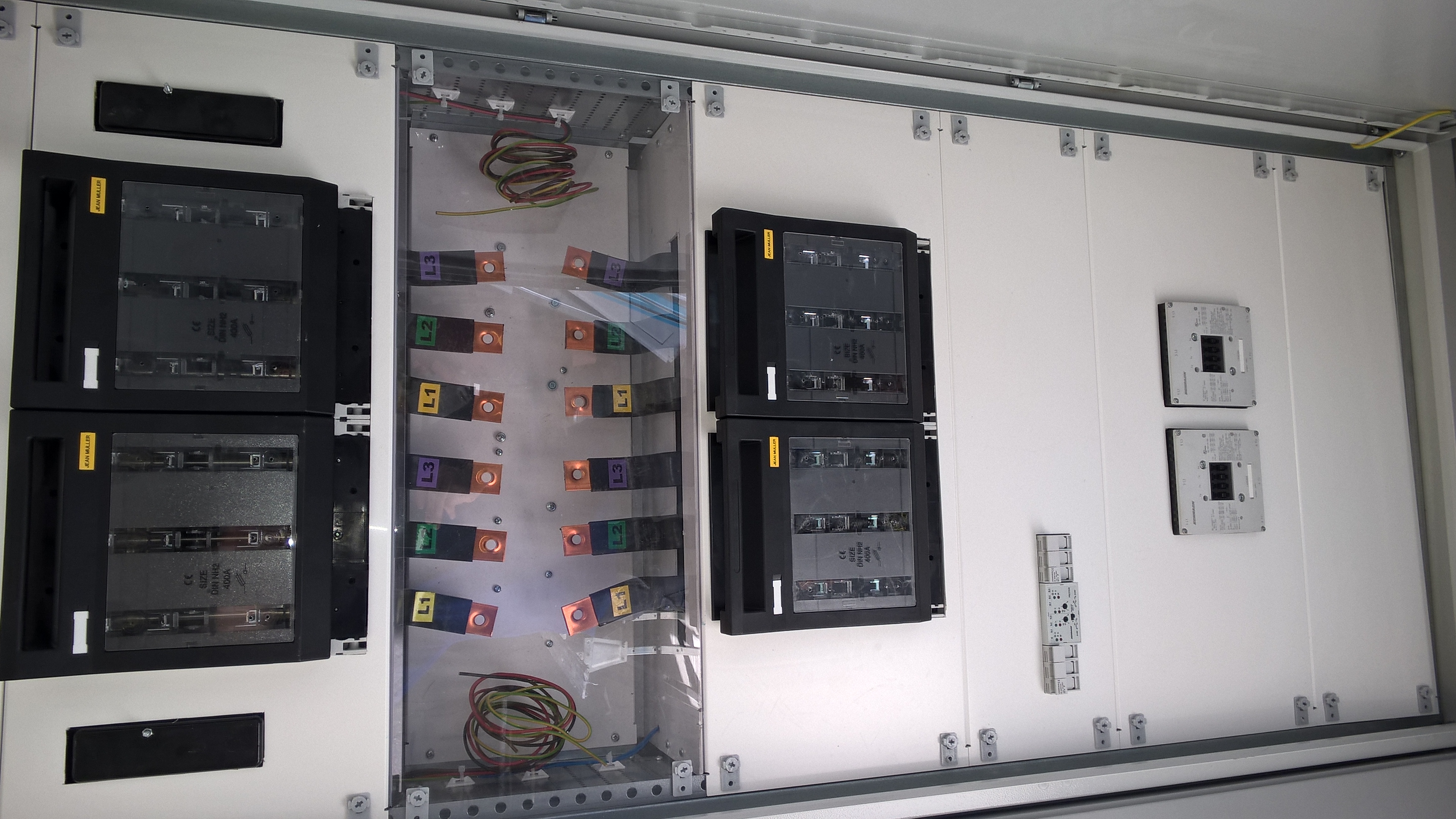 Rozdzielnica elektryczna z bezpiecznikami i przewodami, widoczne oznaczenia faz L1, L2, L3. Elementy zamontowane w białej szafie, widoczne śruby mocujące.