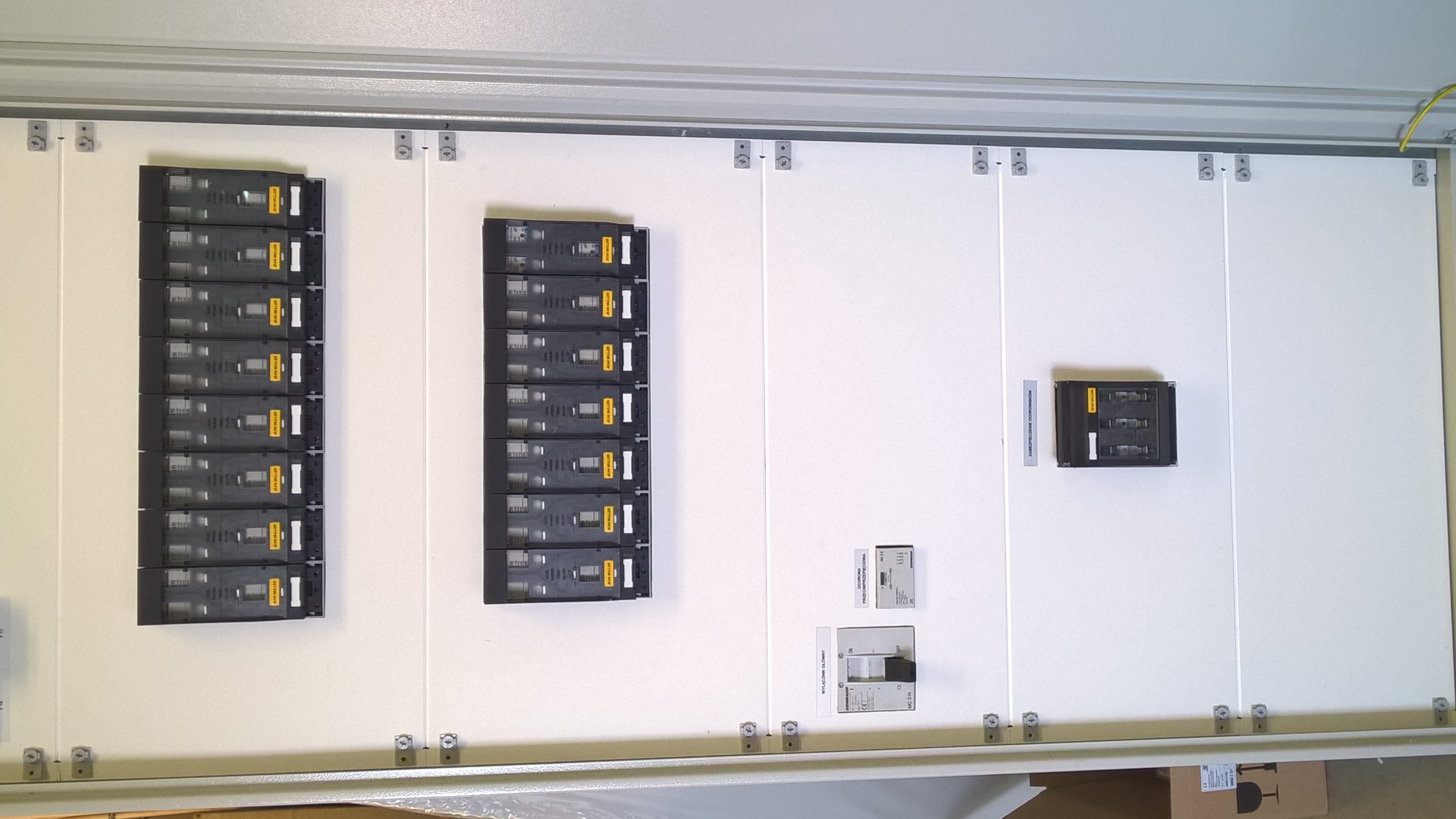 Rozdzielnica elektryczna z zamontowanymi bezpiecznikami i wyłącznikiem głównym, widok z bliska na białą płytę montażową i zainstalowane komponenty.