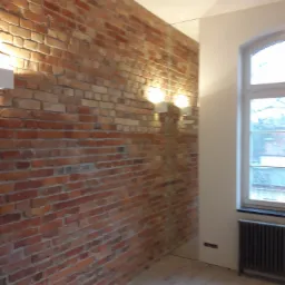 Ceglana ściana w pokoju z oknem, grzejnikiem i drewnianą podłogą. Widoczne lampy ścienne i odbicie ściany w lustrze.