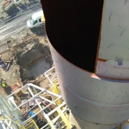 Widok z góry na budowę hali stalowej: fragment konstrukcji z blachy trapezowej, rusztowania, wykop w ziemi i biały samochód dostawczy w tle.