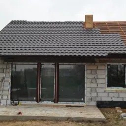 Budowa domu jednorodzinnego w trakcie: wstawione okna, częściowo położony dach z dachówki, obok drabina i auto. Widok na budynek z zewnątrz.