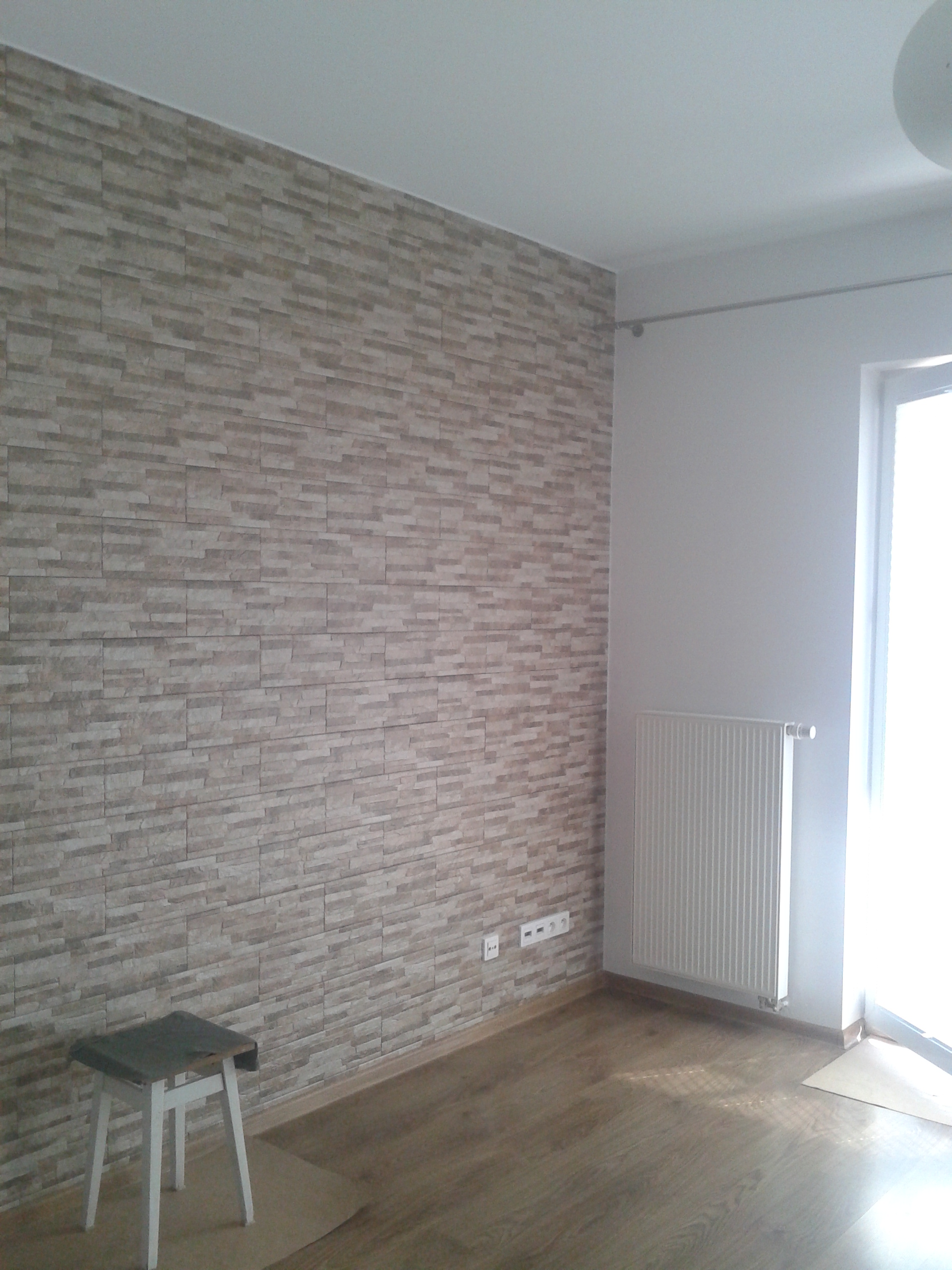 Ściana wykończona dekoracyjną okładziną imitującą kamień, drewniana podłoga, biały grzejnik i fragment białego stołka. Widoczne gniazdka elektryczne.
