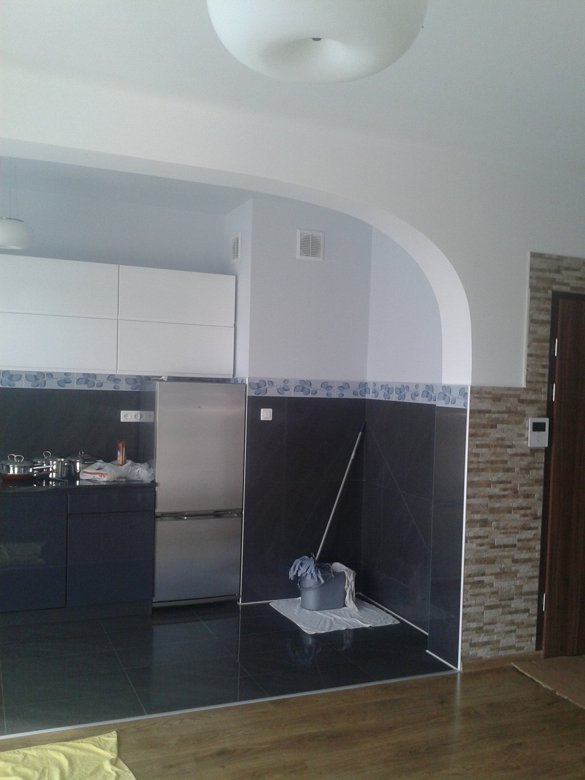 Nowoczesna kuchnia po remoncie: czarne płytki, białe szafki, fragment ściany z imitacją kamienia. Widoczne wiadro z mopem, co sugeruje prace porządkowe po zakończeniu prac.