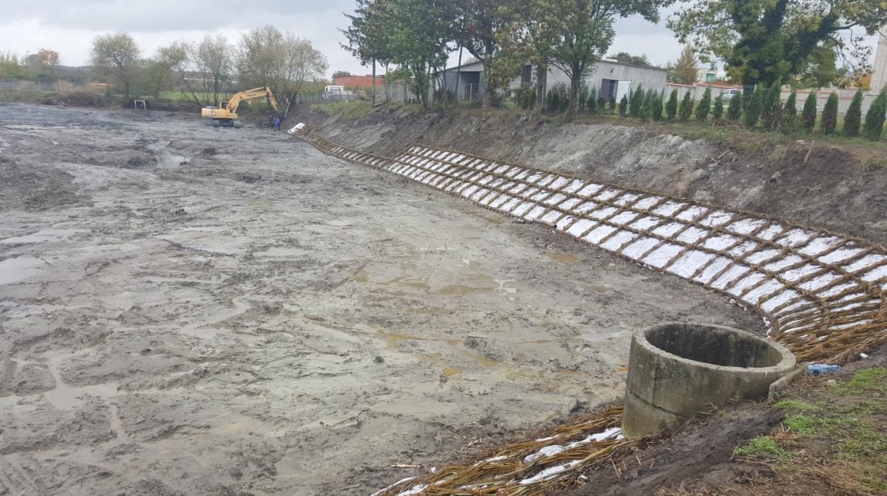 Wykop ziemny z zabezpieczonym brzegiem siatką wiklinową wypełnioną materiałem izolacyjnym, w tle koparka i budynek. Prace ziemne w toku, widoczny betonowy krąg.