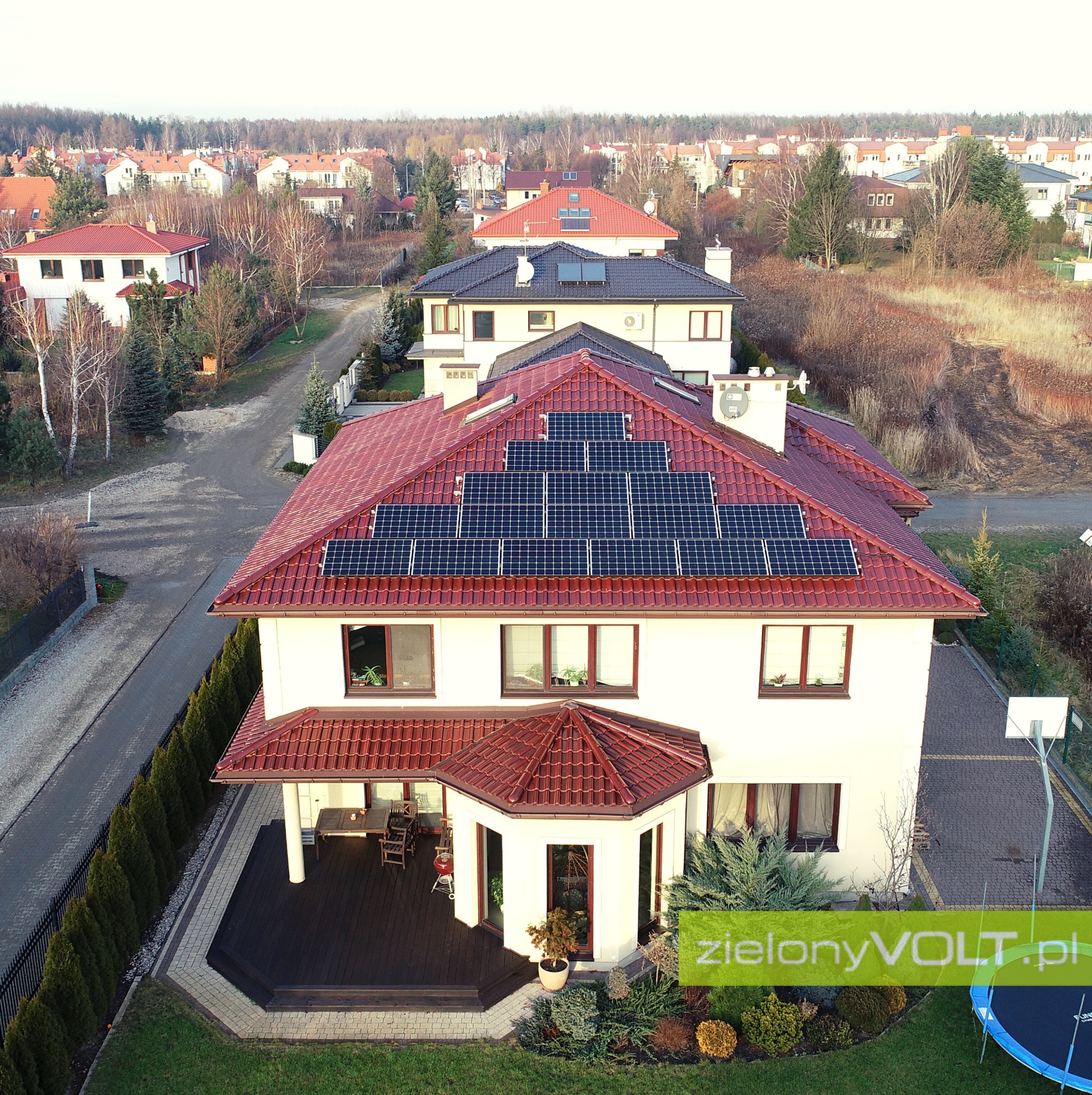 Widok z lotu ptaka na dom jednorodzinny z czerwonym dachem, na którym zamontowane są panele słoneczne; w tle zabudowa osiedlowa.
