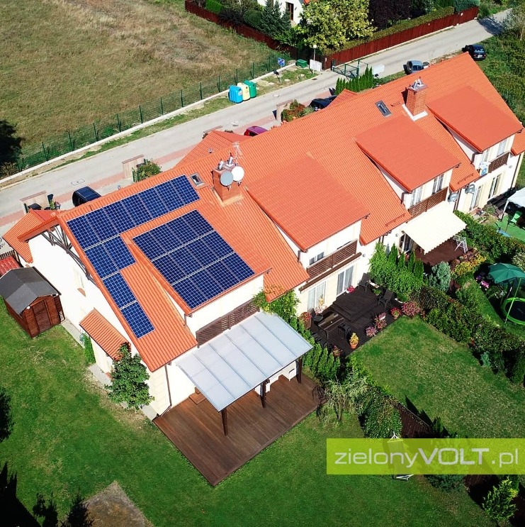 Widok z lotu ptaka na dom z czerwoną dachówką i panelami fotowoltaicznymi na dachu, z widocznym ogrodem i tarasem.