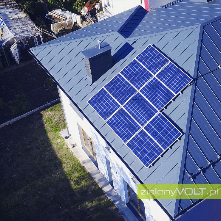 Widok z góry na dom z szarą, metalową dachówką i zainstalowanymi panelami słonecznymi w układzie 3x3 nad kominem.