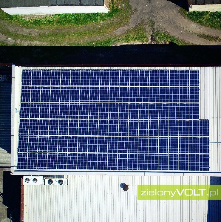 Widok z góry na panele słoneczne zamontowane na dachu budynku przemysłowego, z widocznymi klimatyzatorami i logiem firmy.