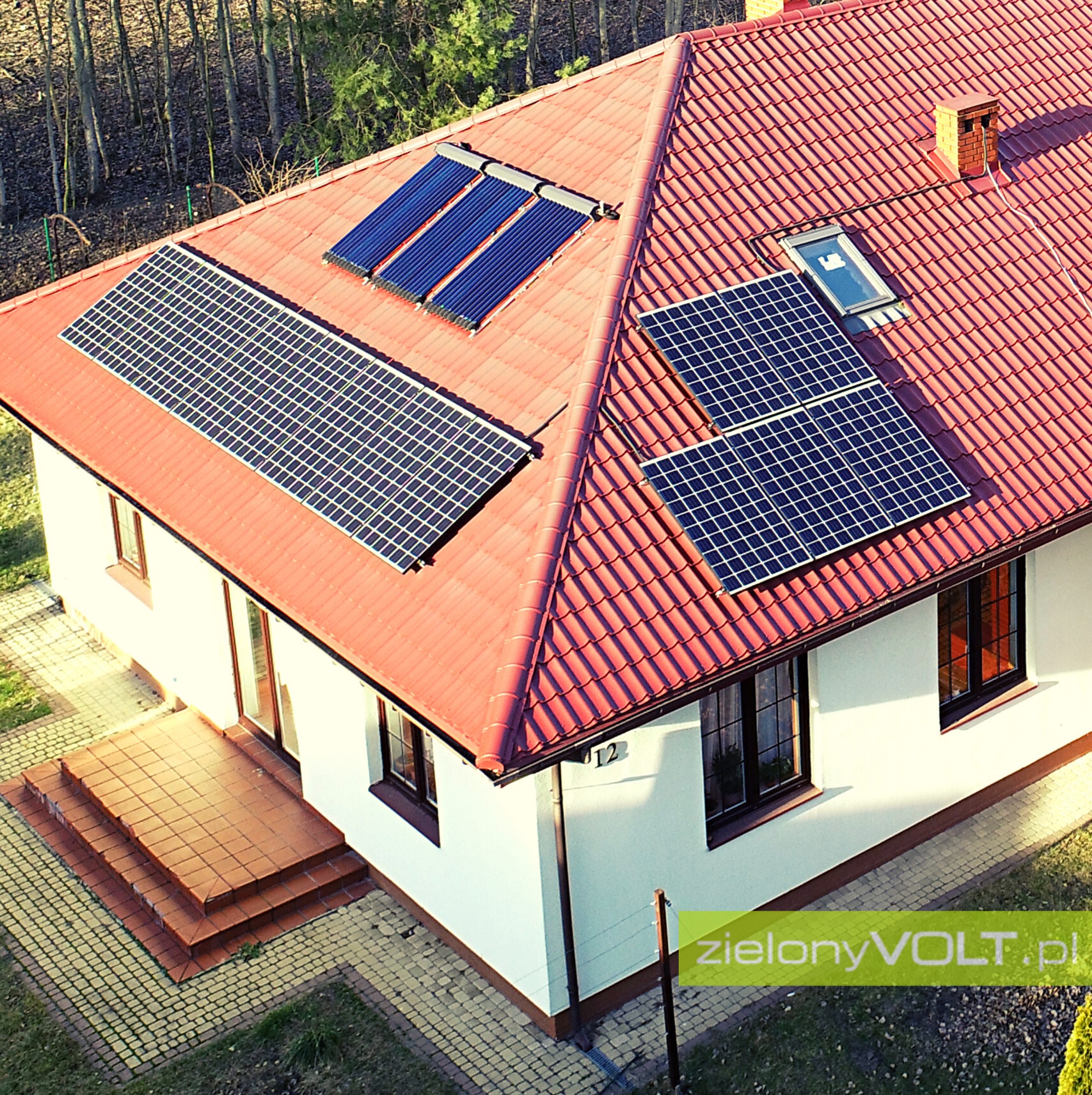 Widok z góry na dom z czerwonym dachem, na którym zamontowano panele słoneczne i kolektory słoneczne; z boku widoczny numer domu '12'.