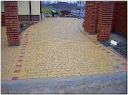 Żółta kostka brukowa ułożona w łukowaty wzór, obrzeża z czerwonej kostki, widoczne słupki z czerwonej cegły i trawnik w tle.