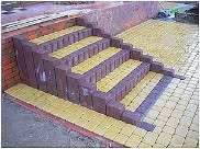 Schody z kostki brukowej w odcieniach brązu i żółci, prowadzące do wyżej położonego terenu. Widoczne detale ułożenia i wykończenia.