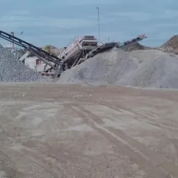 Widok na plac budowy z przesiewaczem i hałdami kruszywa o różnej frakcji, widoczne ślady opon pojazdów transportowych.