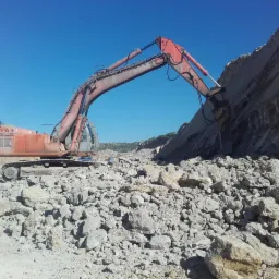 Czerwony koparko-ładowarka z pneumatycznym młotem rozbija skalną ścianę w kamieniołomie, z dużą ilością gruzu na pierwszym planie i błękitnym niebem w tle.