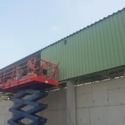 Pracownicy na podnośniku montują blaszane elementy dachu na betonowej konstrukcji budynku w słoneczny dzień. Widoczna metalowa konstrukcja dachu.