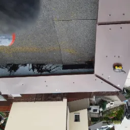 Widok z góry na fragment dachu pokrytego papą z brązowymi obróbkami blacharskimi, widoczna zwinięta miarka budowlana na obróbce.