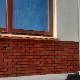 Fragment elewacji budynku z oknami zabezpieczonymi folią, wykończony czerwoną cegłą klinkierową, widoczny pracownik z wiadrem w trakcie prac wykończeniowych.