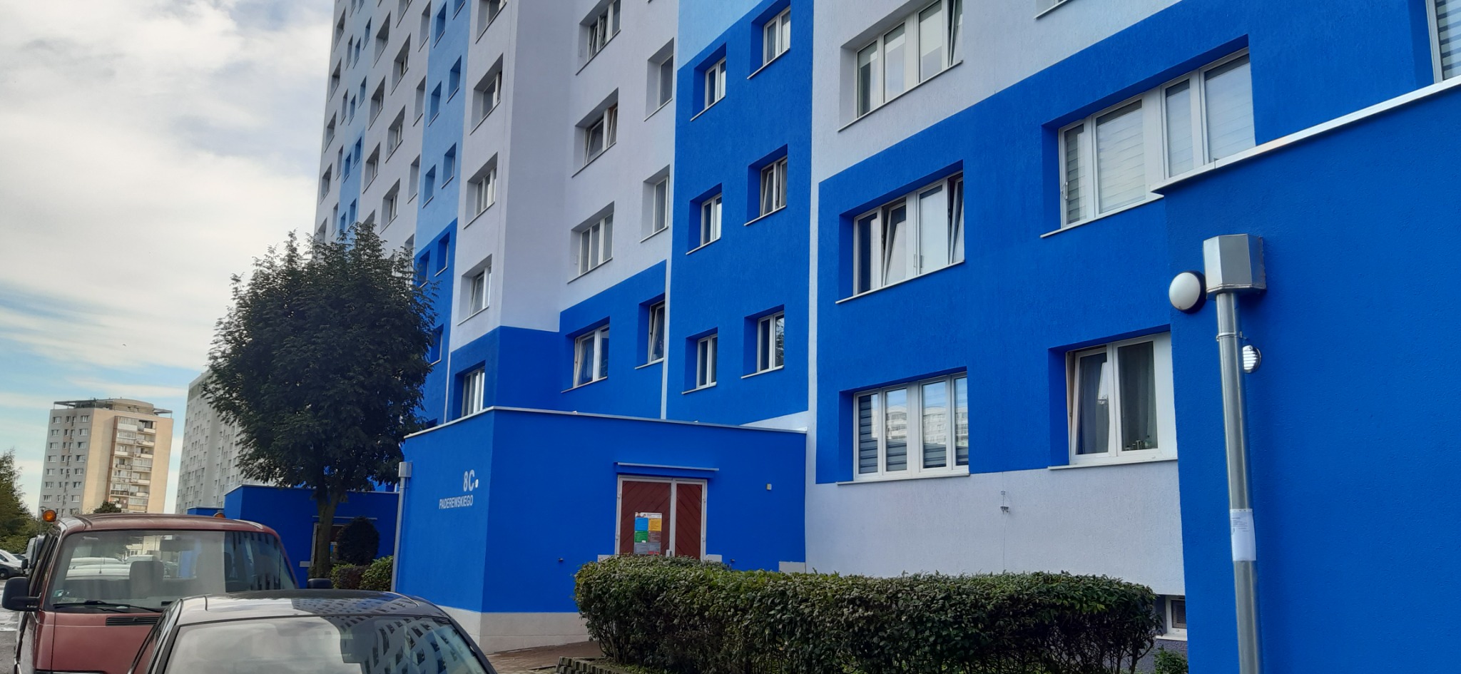 Odnowiony blok mieszkalny z elewacją w odcieniach błękitu i szarości, widoczny z poziomu ulicy, z zaparkowanymi samochodami w tle.